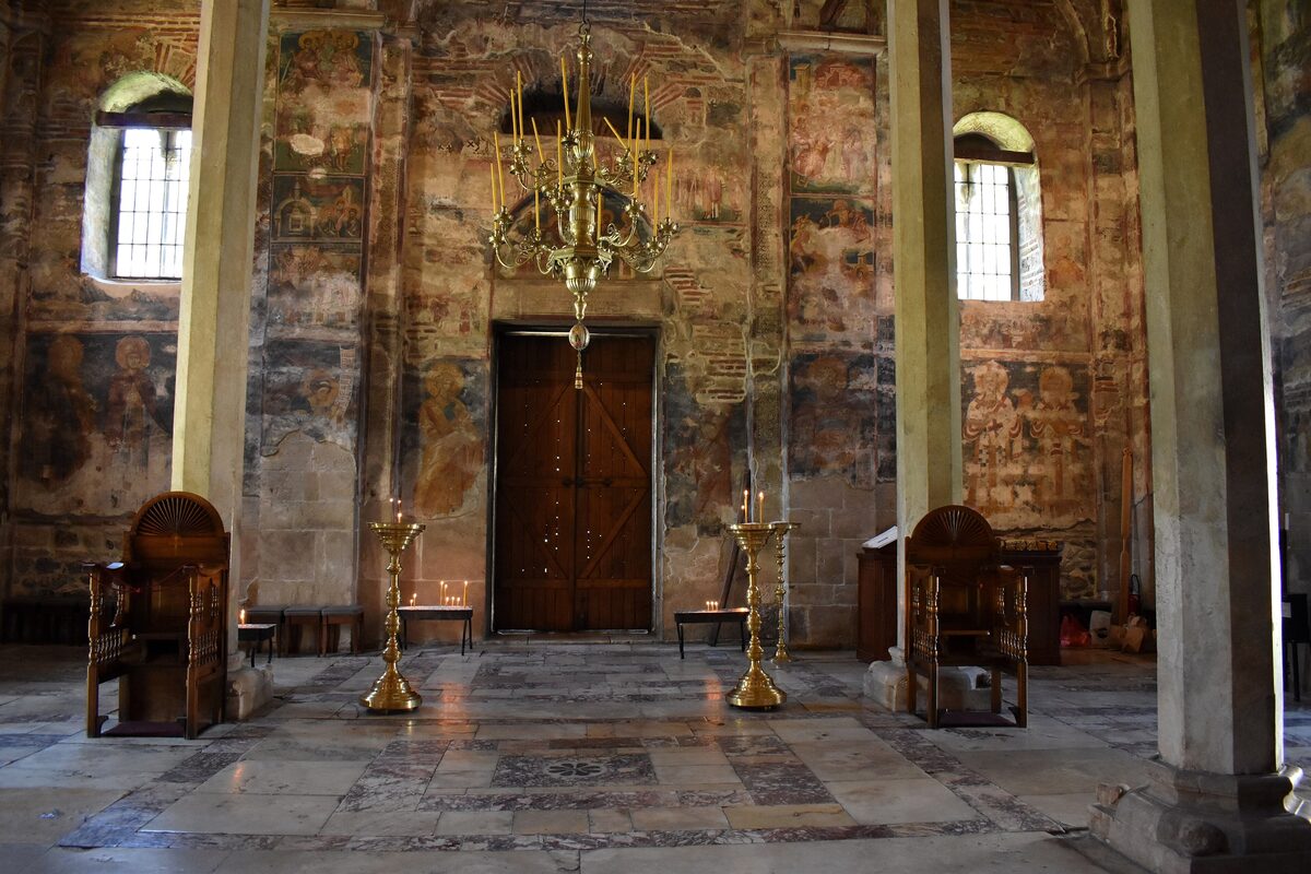 Inside the Visoki Dečani church: fresco-covered walls, slender stone columns, a brass chandelier and carved wooden thrones around a patterned marble floor.