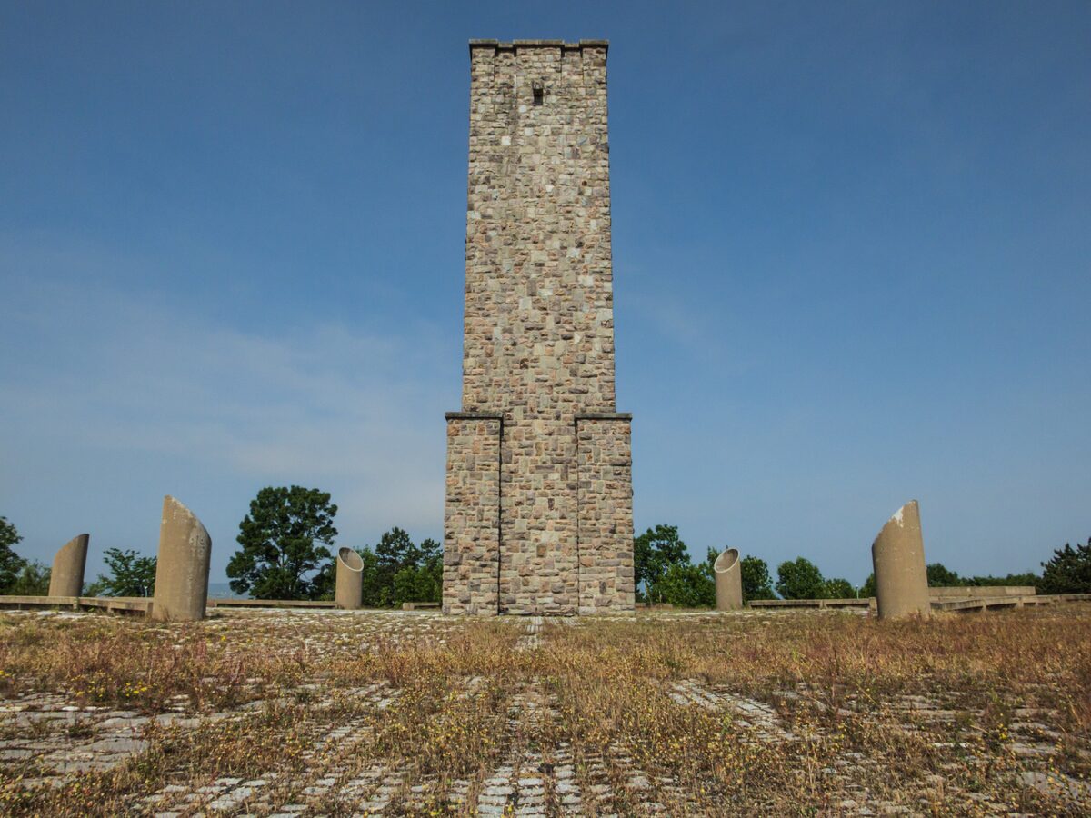 The stone tower of the Gazimestan monument under a clear blue sky, ringed by cannon-barrel sculptures on a grassy paved plateau.