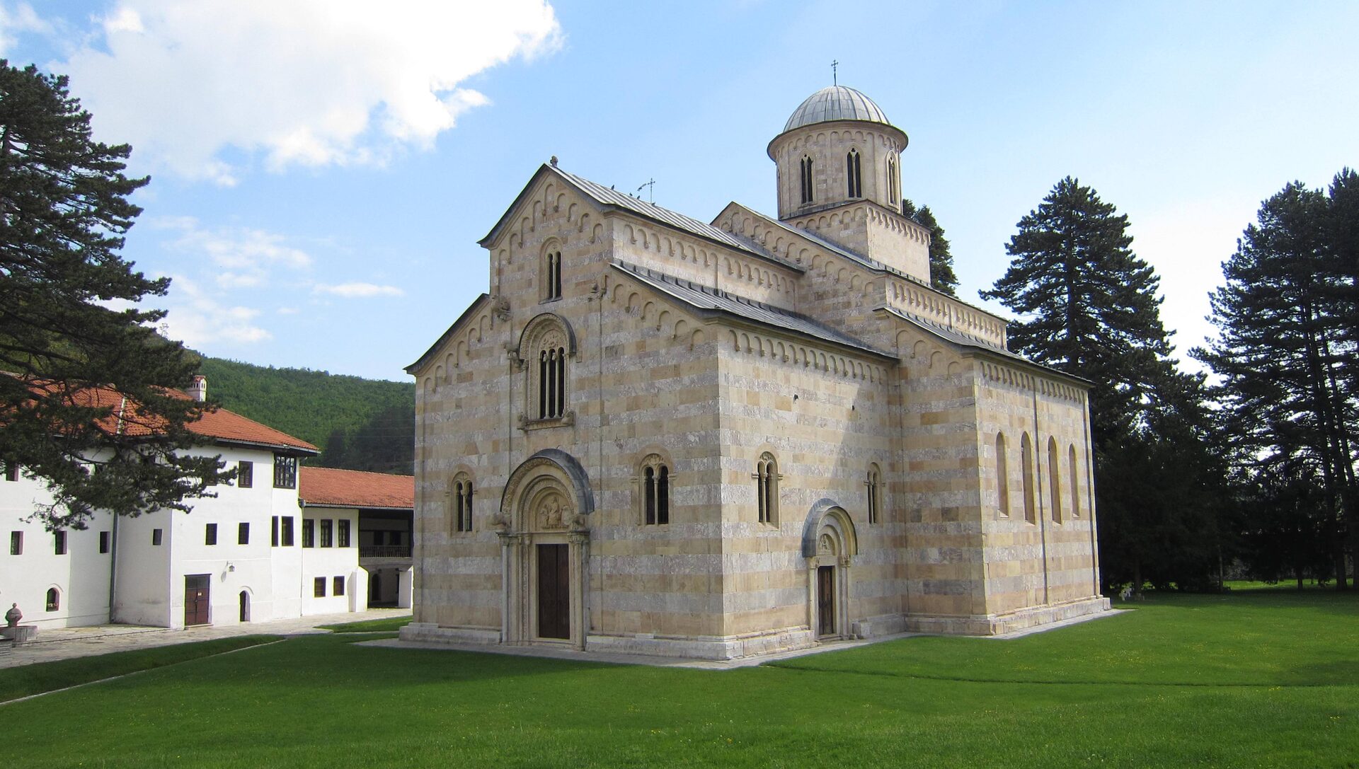The striped marble Church of Christ Pantocrator at Visoki Dečani on its green lawn, with the white monastery lodgings, tall pines and a wooded hillside behind.