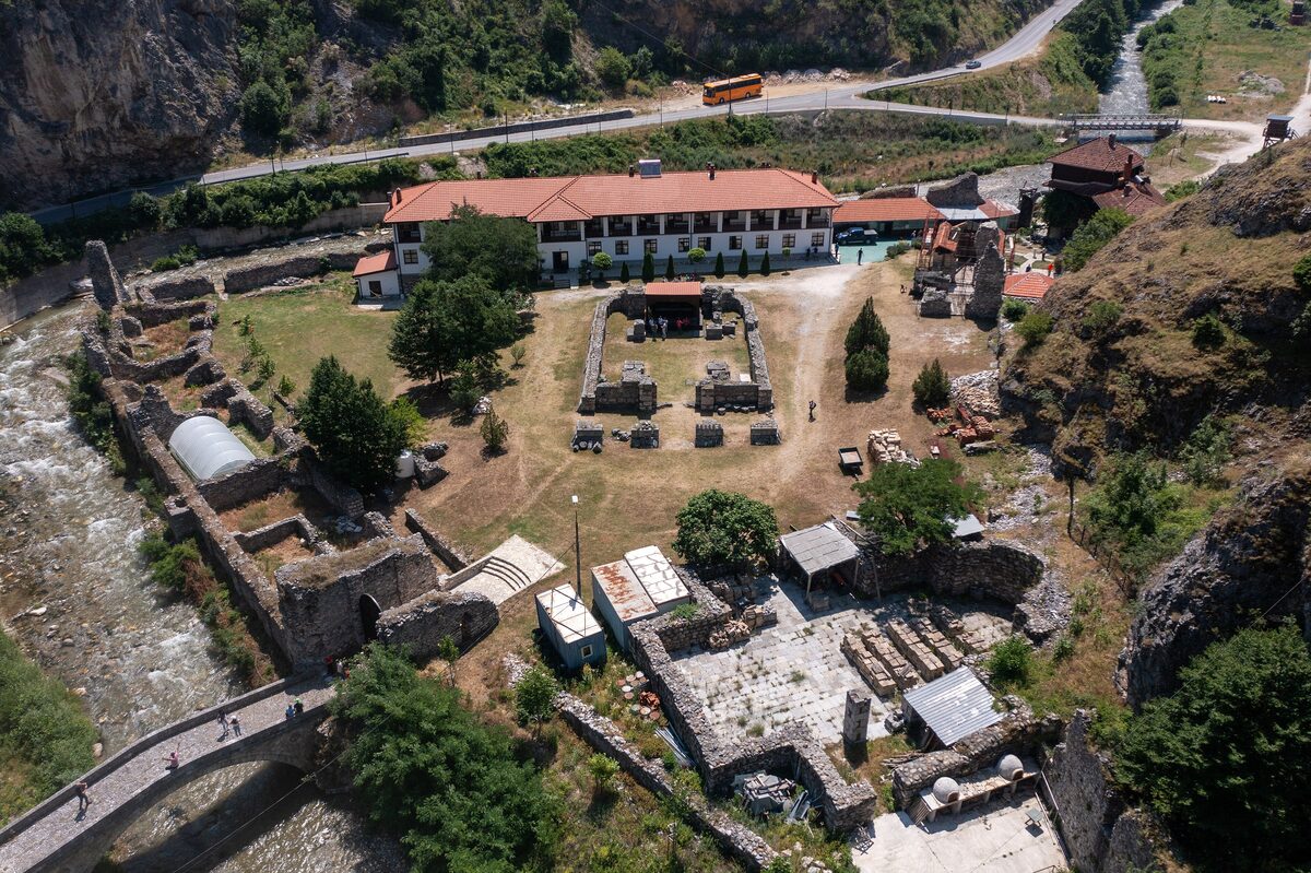 Aerial view of the Holy Archangels monastery near Prizren: excavated church foundations and ruined walls beside the Bistrica river and a stone footbridge, with the new monastery lodgings and the gorge road behind.