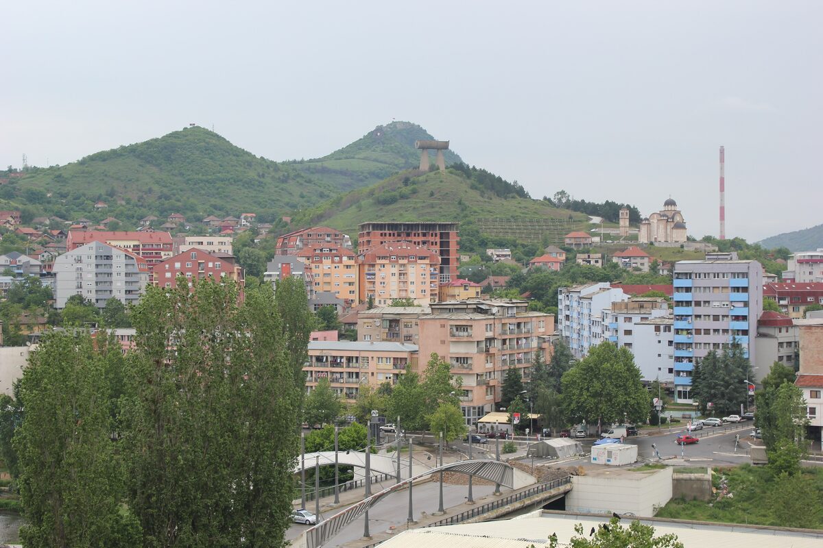 North Kosovska Mitrovica seen from above, with the arched Ibar bridge in front, apartment blocks, St Demetrius church on the hill to the right and the Miners monument on the green hill behind.