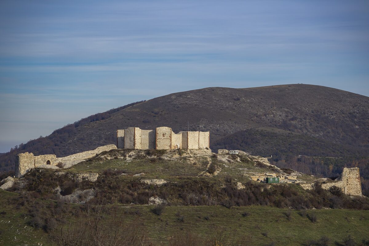 The ruined towers and curtain walls of Novo Brdo fortress on a grassy hilltop, with a bare mountain rising behind.