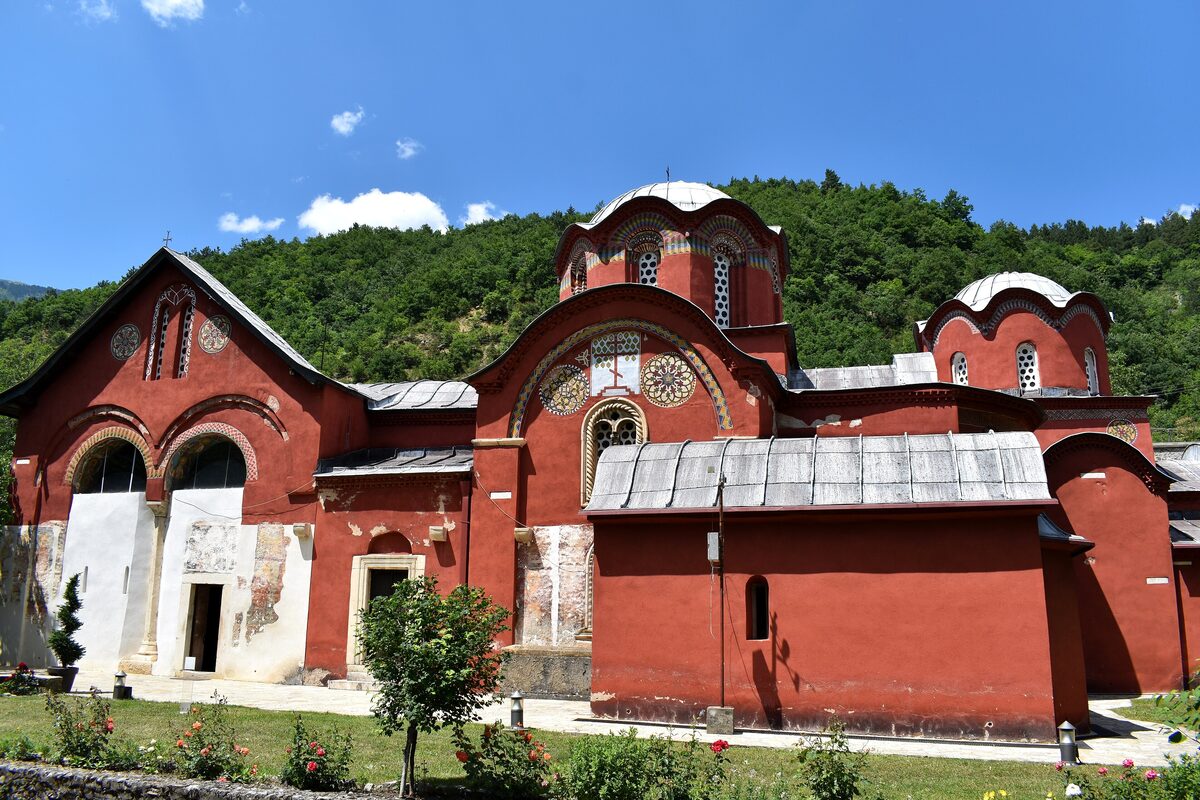 The red-painted churches of the Patriarchate of Peć with their domes and arched narthex, against a forested hillside and blue summer sky.
