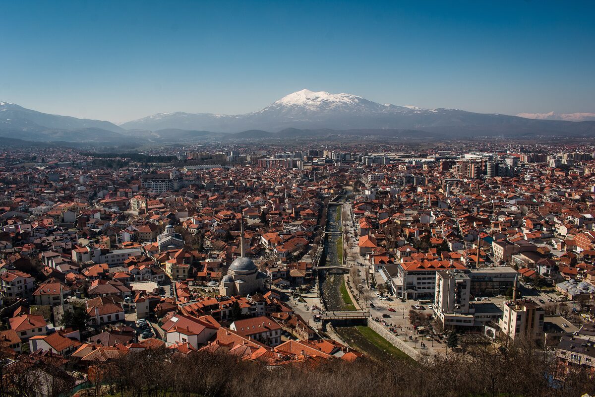 Prizren seen from the fortress: a sea of red-tiled roofs, the dome and minaret of the Sinan Pasha mosque and the Bistrica river running through the centre, with a snow-capped mountain beyond.