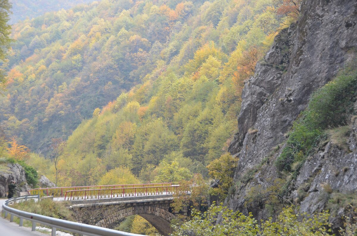 The Rugova gorge road crossing a stone-arched bridge beneath a grey cliff, with steep slopes of autumn forest in yellow and orange behind.