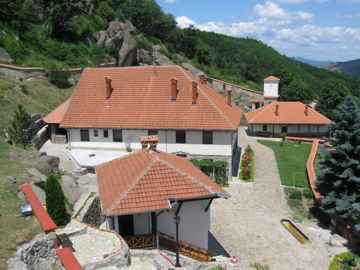 The Sokolica monastery complex from above: red-tiled lodgings, a flower-lined stone path and a small bell tower, set among granite boulders on a wooded hillside.
