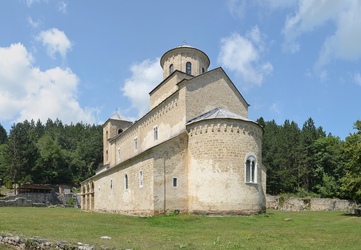 The pale stone Church of the Holy Trinity at Sopoćani monastery seen from the east, with its apse, dome and bell tower, in a meadow ringed by pine forest.