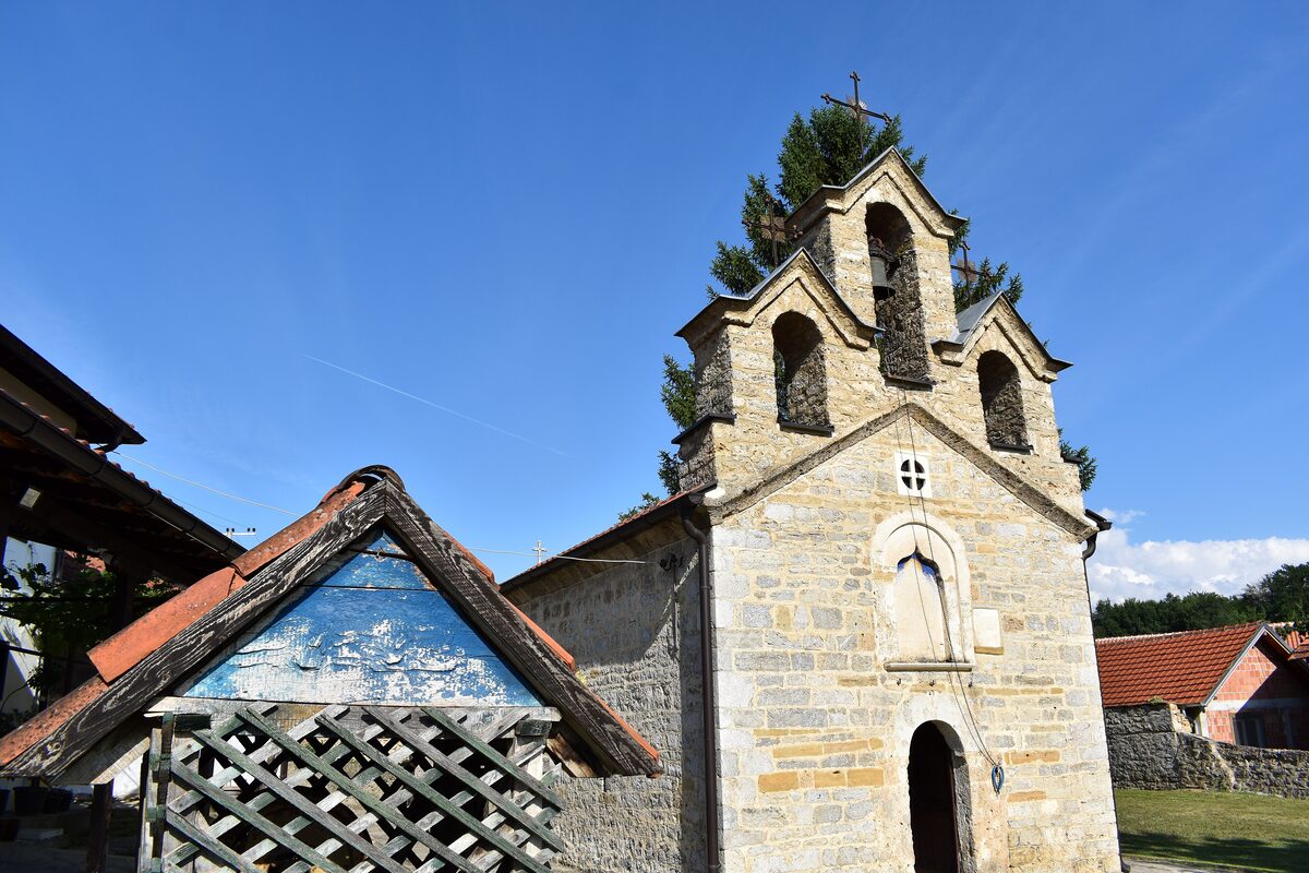 The stone Church of St Stephen in Velika Hoča with its triple bell gable, beside a wooden wellhead under a clear blue sky.