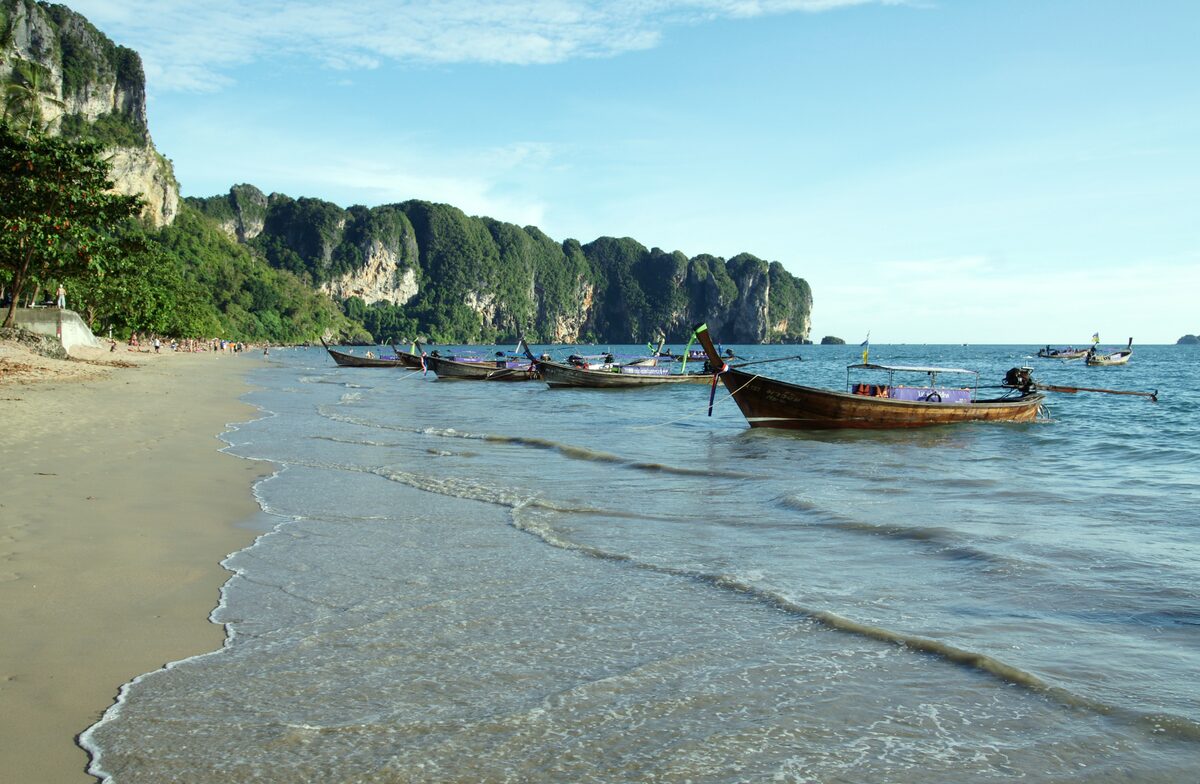 Longtail boats on the sand at Ao Nang beach in sunshine, with limestone cliffs at the far end of the bay