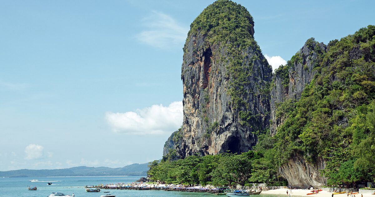 Limestone cliffs above Phra Nang beach in bright sun, with longtail boats moored along the sand and swimmers in the shallows