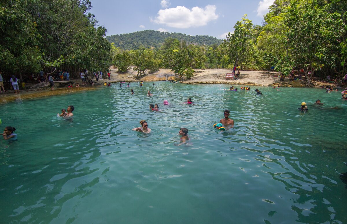 Swimmers in the turquoise Emerald Pool, with forest and sandy banks around the water