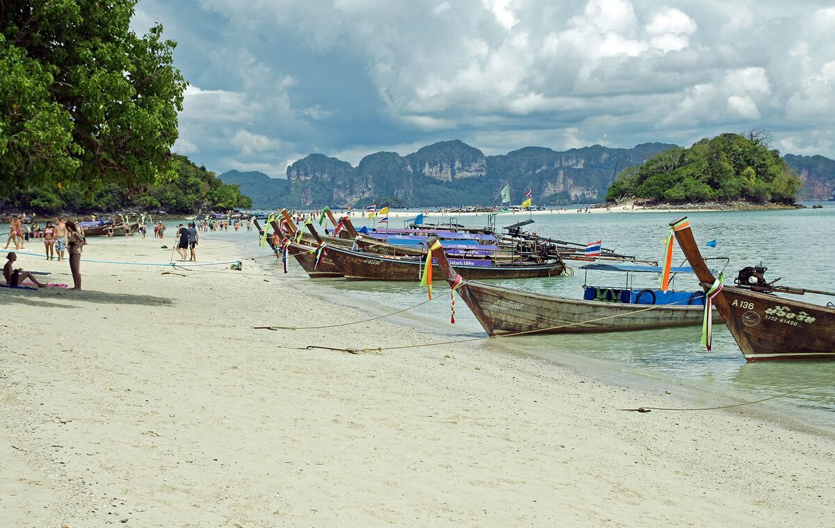 Longtail boats moored along the white sand of Ko Kai beach, with limestone hills across the water