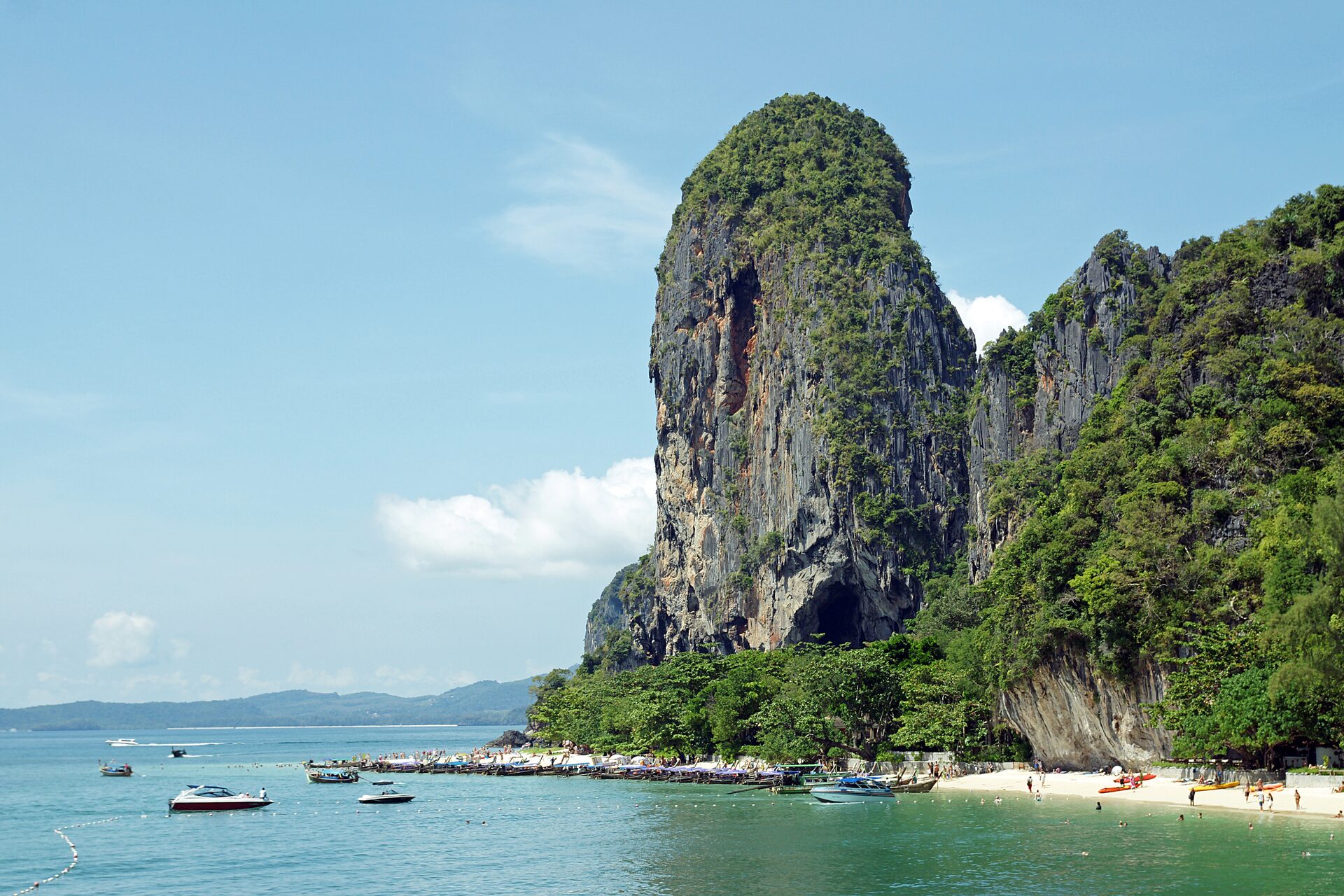 Limestone cliffs above Phra Nang beach in bright sun, with longtail boats moored along the sand and swimmers in the shallows