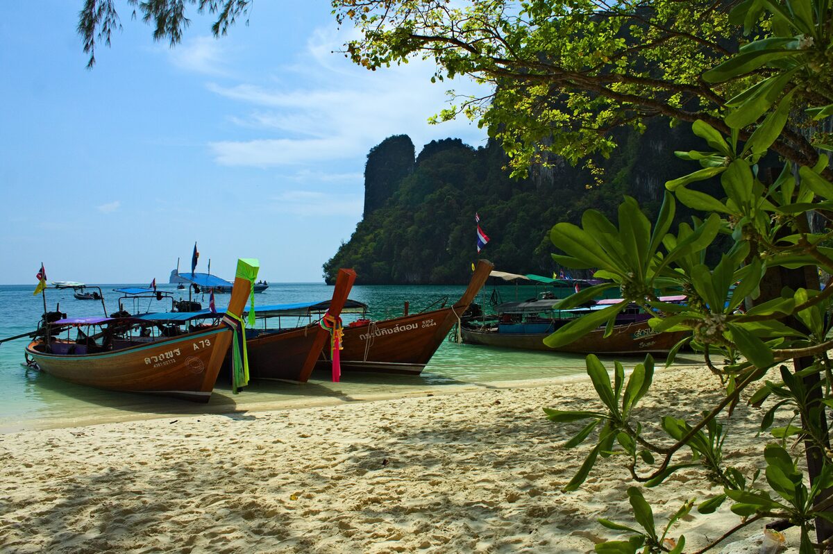 Longtail boats moored on a sandy beach at Koh Hong, with a limestone headland behind and branches framing the shot