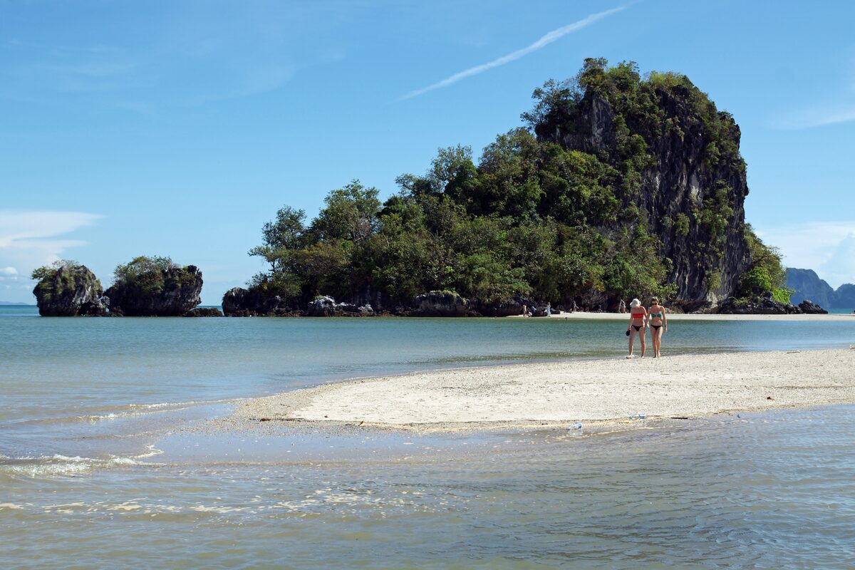 A white sandbar running out from Hat Noppharat Thara beach toward a wooded limestone island, with two people standing on the sand