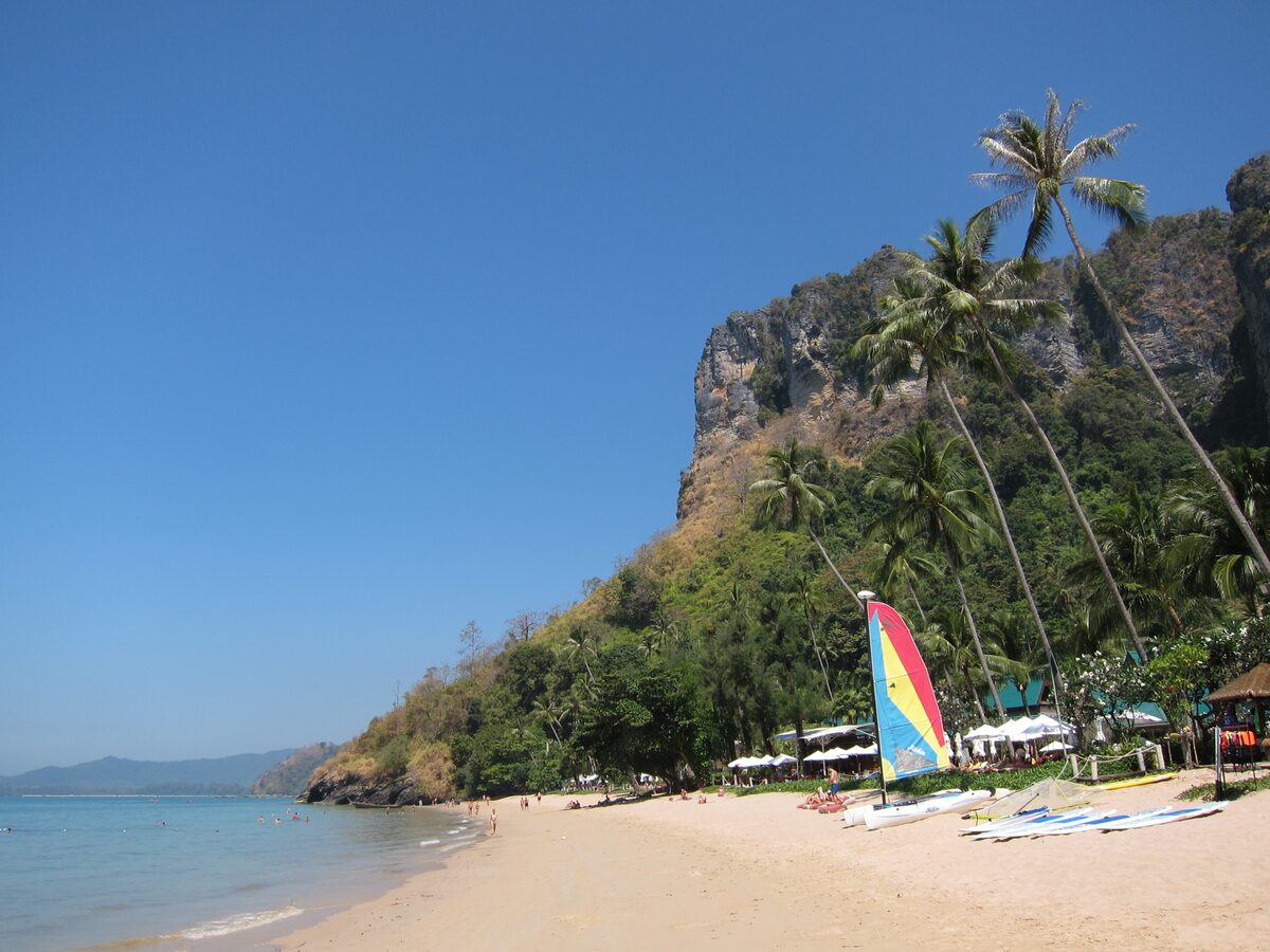 Pai Plong beach on a clear day: pale sand, palms and a forested limestone slope above a calm sea, with a colorful sail on the shore