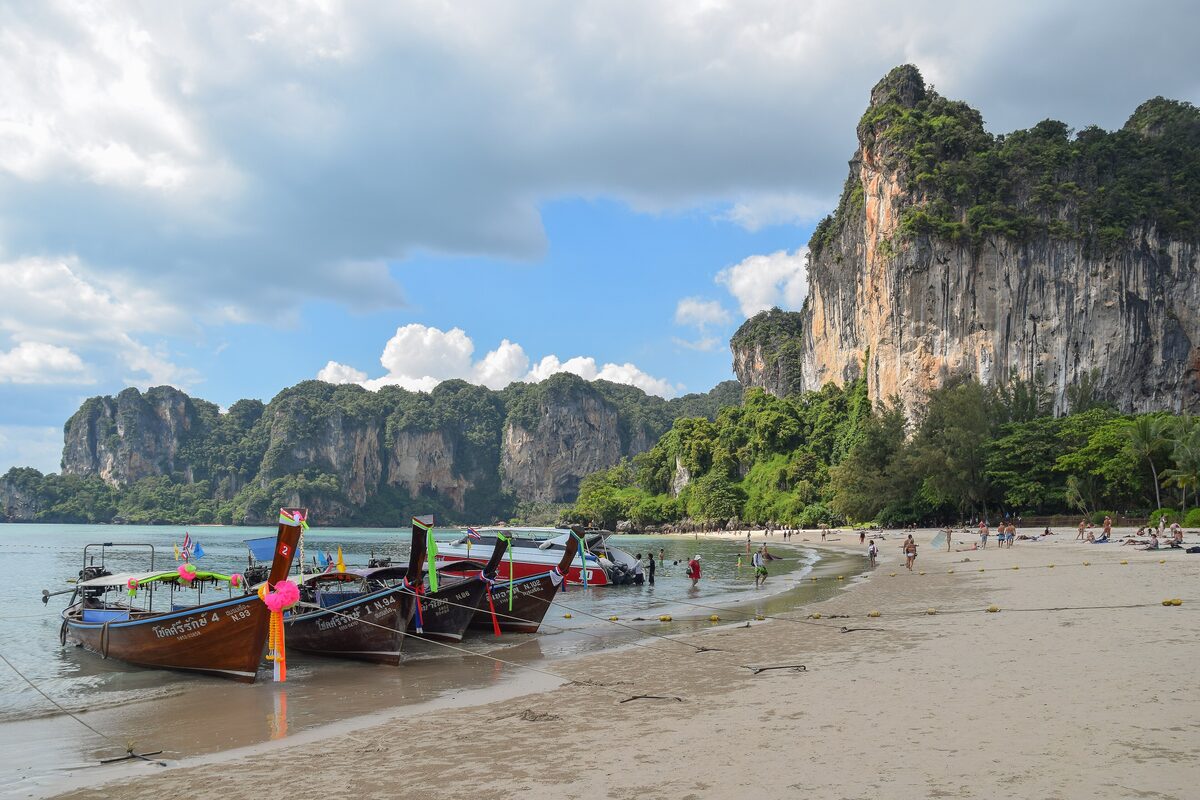 Longtail boats pulled up on the sand at Railay West under a blue sky, with a tall limestone cliff at the end of the beach