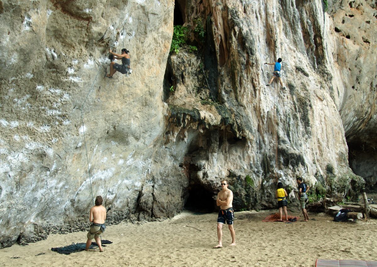 Two climbers on a tall limestone wall above the sand at Phra Nang beach, Railay, with belayers and a cave mouth at the base