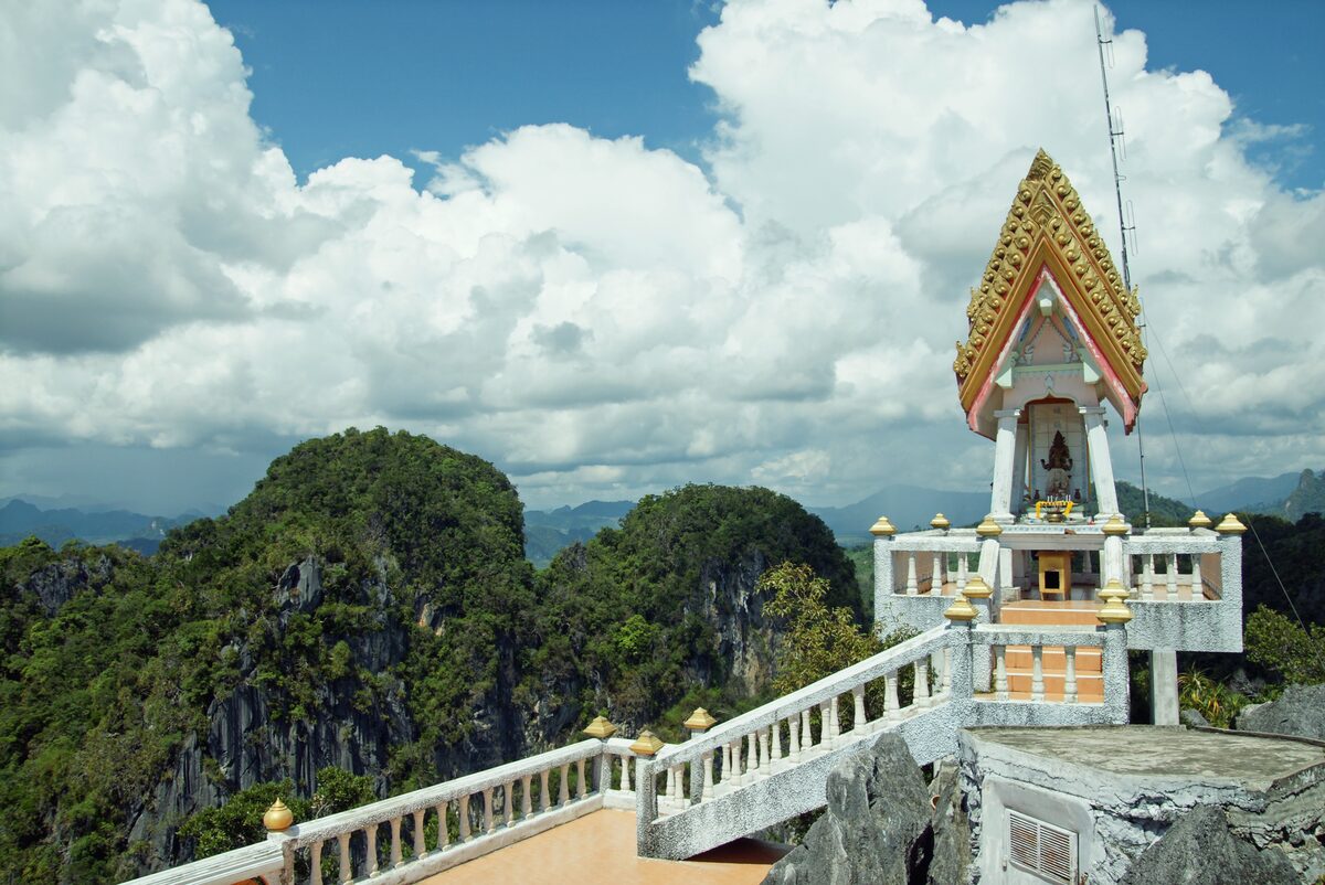Small spired shrine on a white balustraded terrace on the summit of Tiger Cave Temple, with forested karst hills and big clouds behind