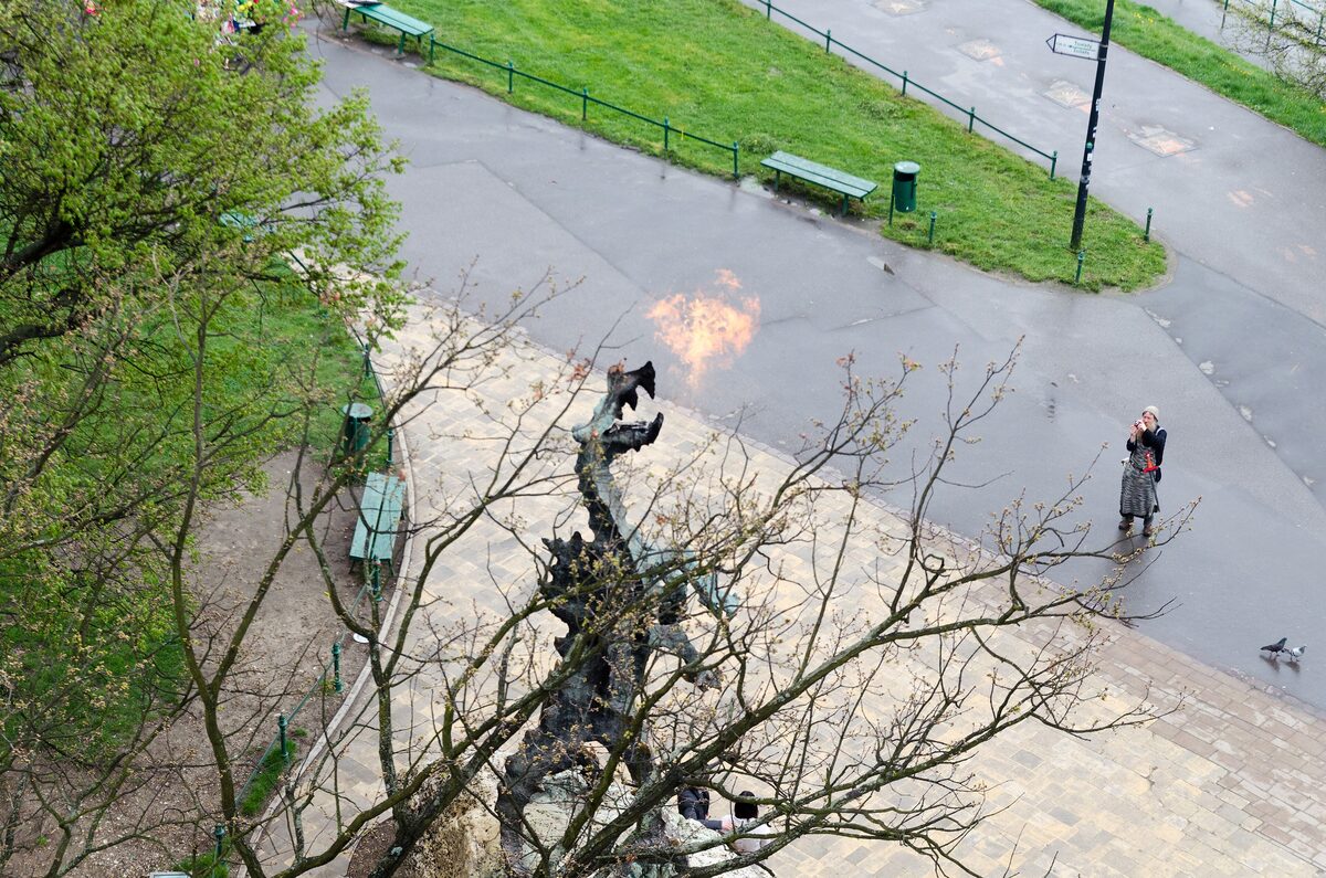 The bronze Wawel Dragon statue breathing a burst of fire on the riverbank below the castle, seen from above