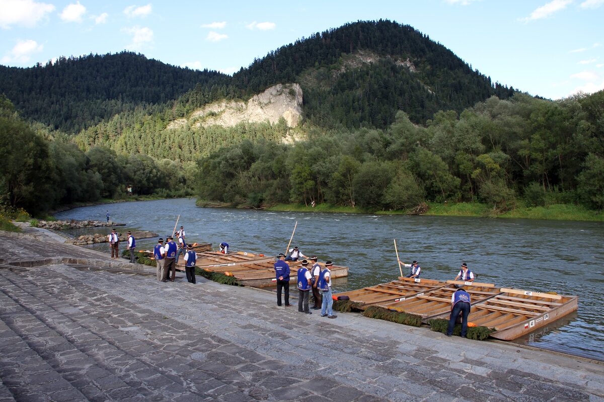 Wooden rafts and raftsmen at a landing on the Dunajec river, with forested hills and a limestone cliff behind