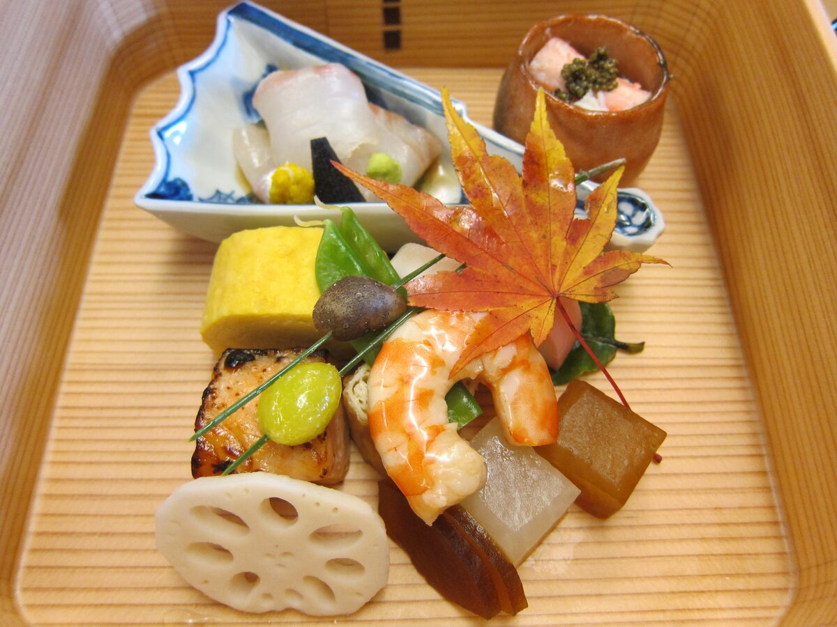 A wooden tray of kaiseki dishes with prawn, lotus root, egg roll and a red maple leaf as garnish