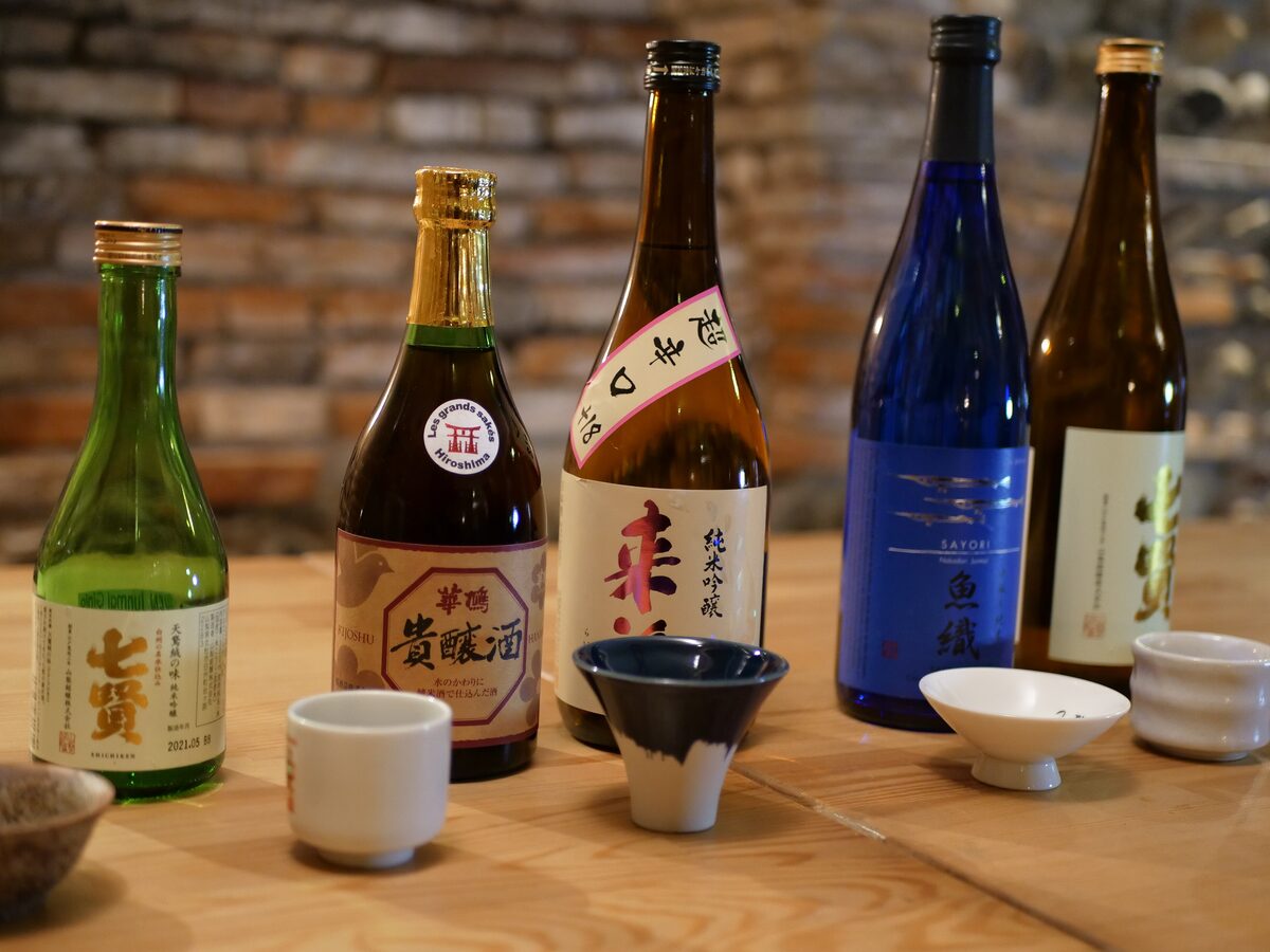 Five bottles of Japanese sake standing on a wooden counter, with small cups in front