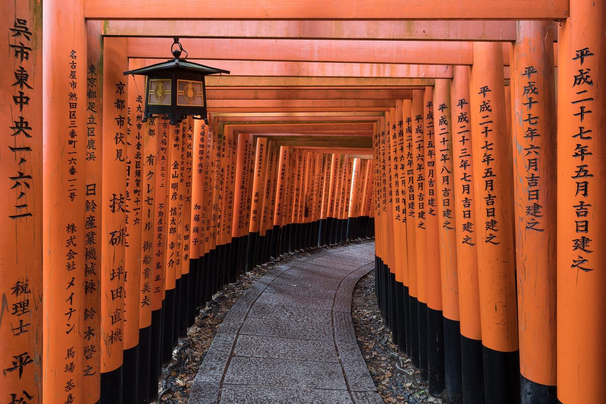 A tunnel of vermilion torii gates on a stone path at Fushimi Inari, with a lantern hanging from the top of one gate