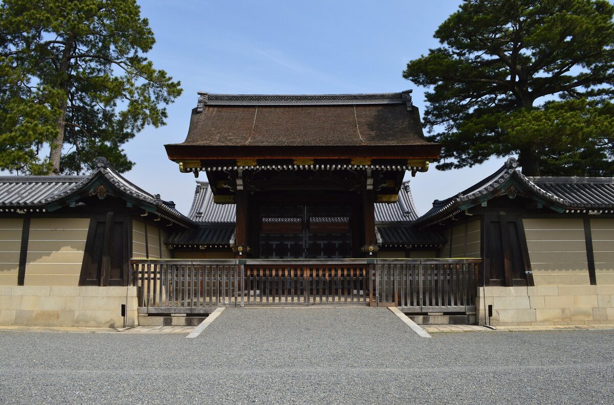 The Kenreimon, the main gate of the Kyoto Imperial Palace, with its dark cypress-bark roof between pine trees and a plastered wall under a blue sky