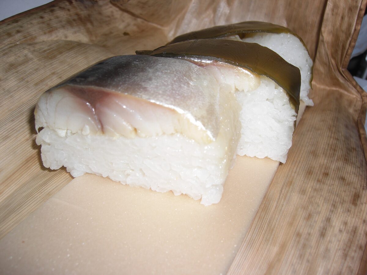 Two thick slices of pressed mackerel sushi, topped with a strip of kelp, lying on a bamboo leaf