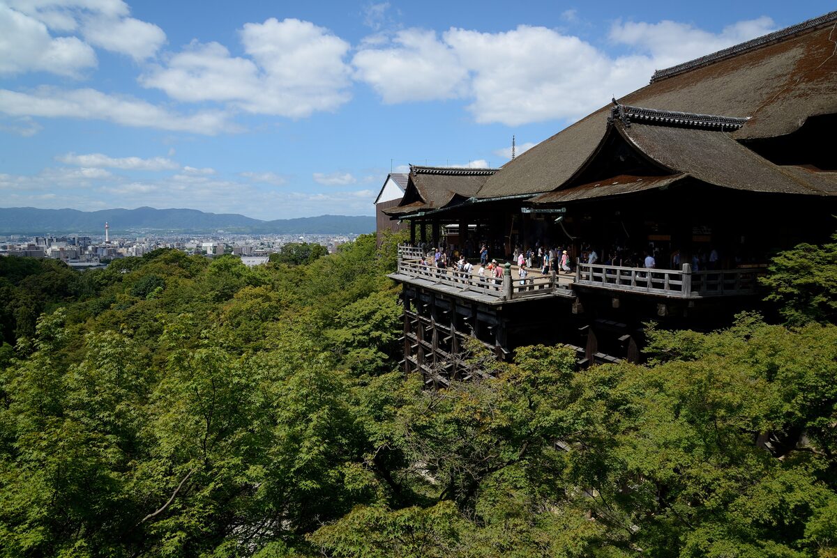 The wooden stage of Kiyomizu-dera jutting out over a sea of green summer trees, with the city of Kyoto and the hills beyond under a blue sky