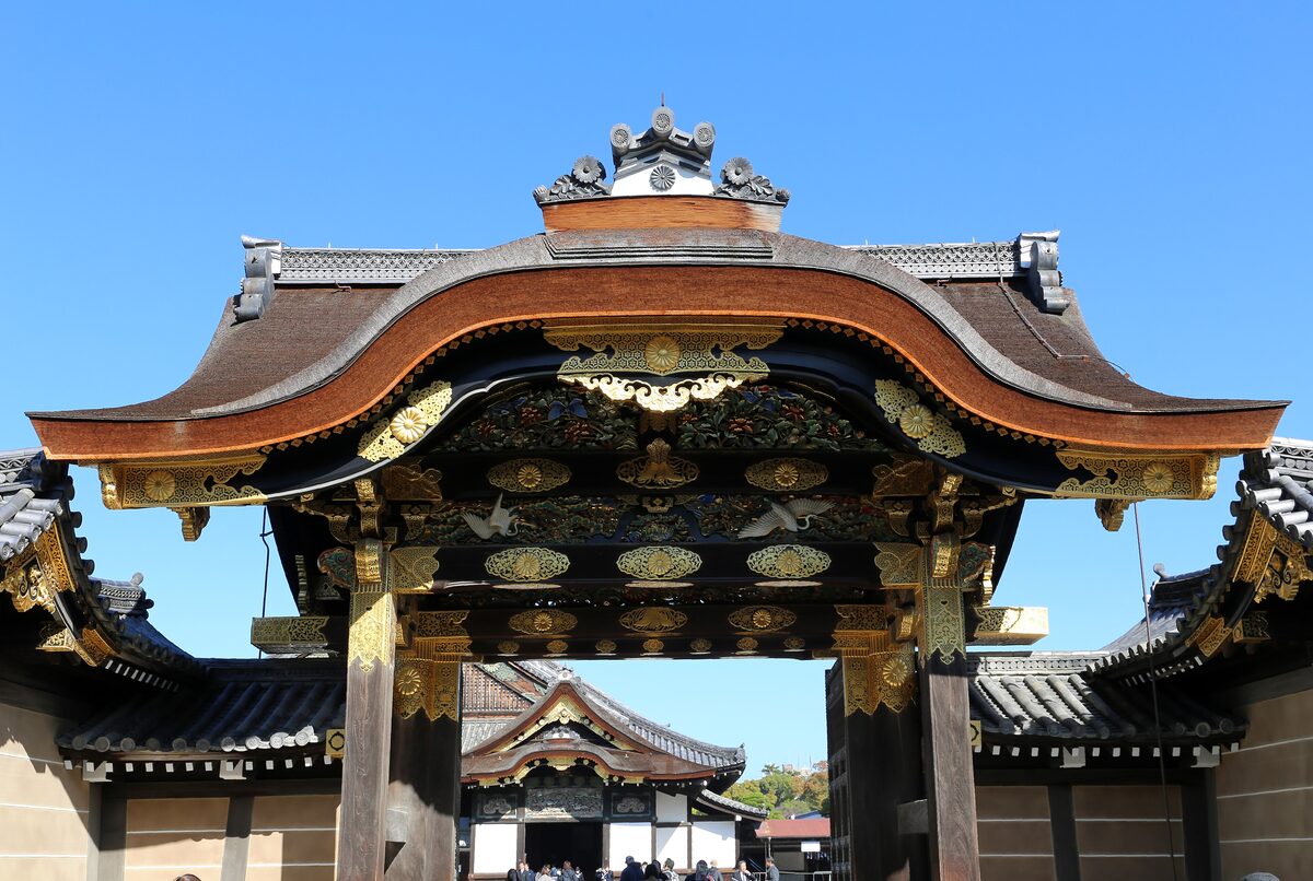 The Karamon gate of Nijo Castle, its curved roof and black-and-gold carvings against a blue sky, with the palace roof glimpsed through the opening