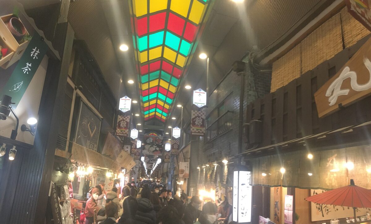 The covered lane of Nishiki Market in the evening, with a colorful striped roof, hanging lanterns and a crowd of shoppers