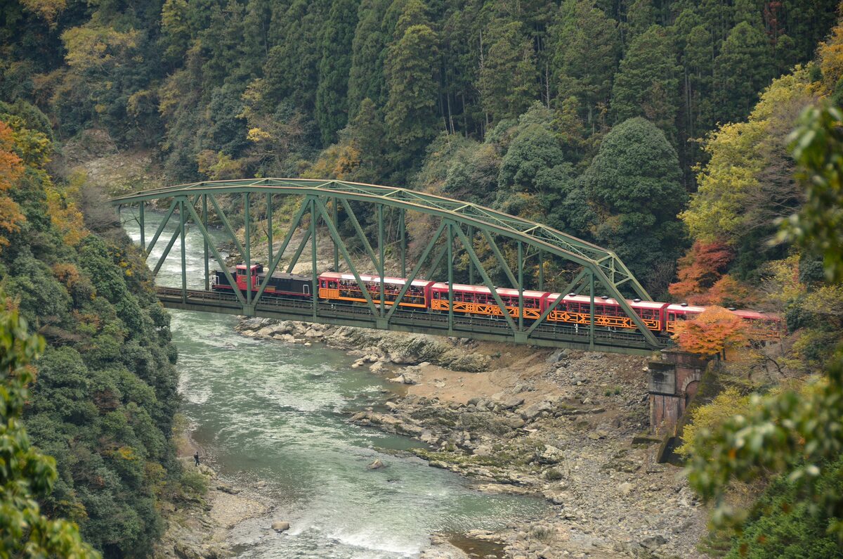 The orange and red Sagano Scenic Railway train crossing a green iron bridge over the Hozu River gorge, with autumn colour on the slopes