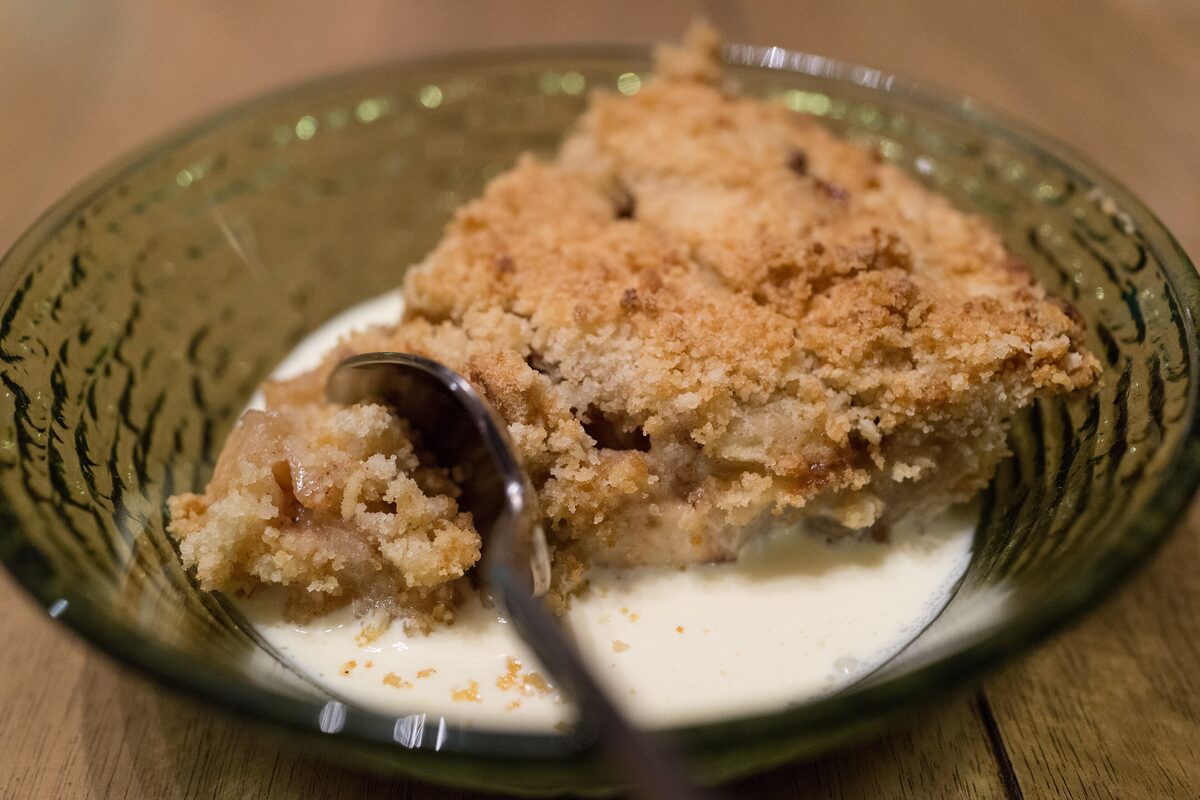 A bowl of crumb-topped apple pie with a spoon and a pour of cream