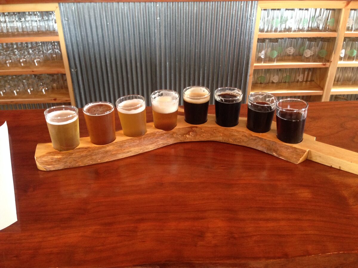 A flight of eight beers in small glasses on a wooden paddle along a bar