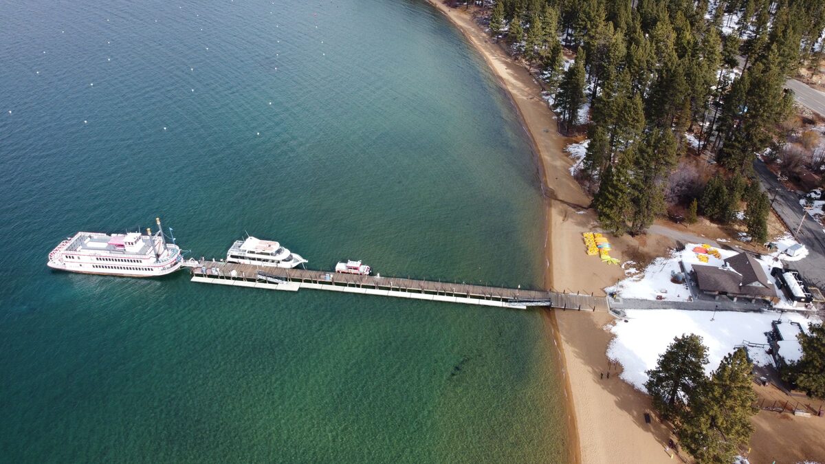 Two cruise boats at the long pier at Zephyr Cove, on Lake Tahoe's Nevada shore in winter, seen from above; the Spirit of Tahoe sails from Ski Run Marina