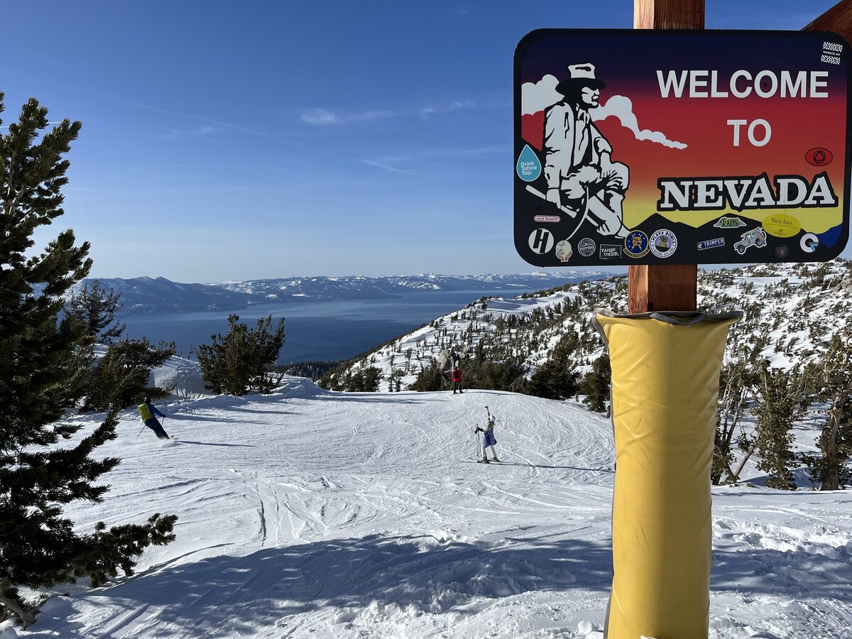 A Welcome to Nevada sign at the top of a snowy Heavenly run, with Lake Tahoe below