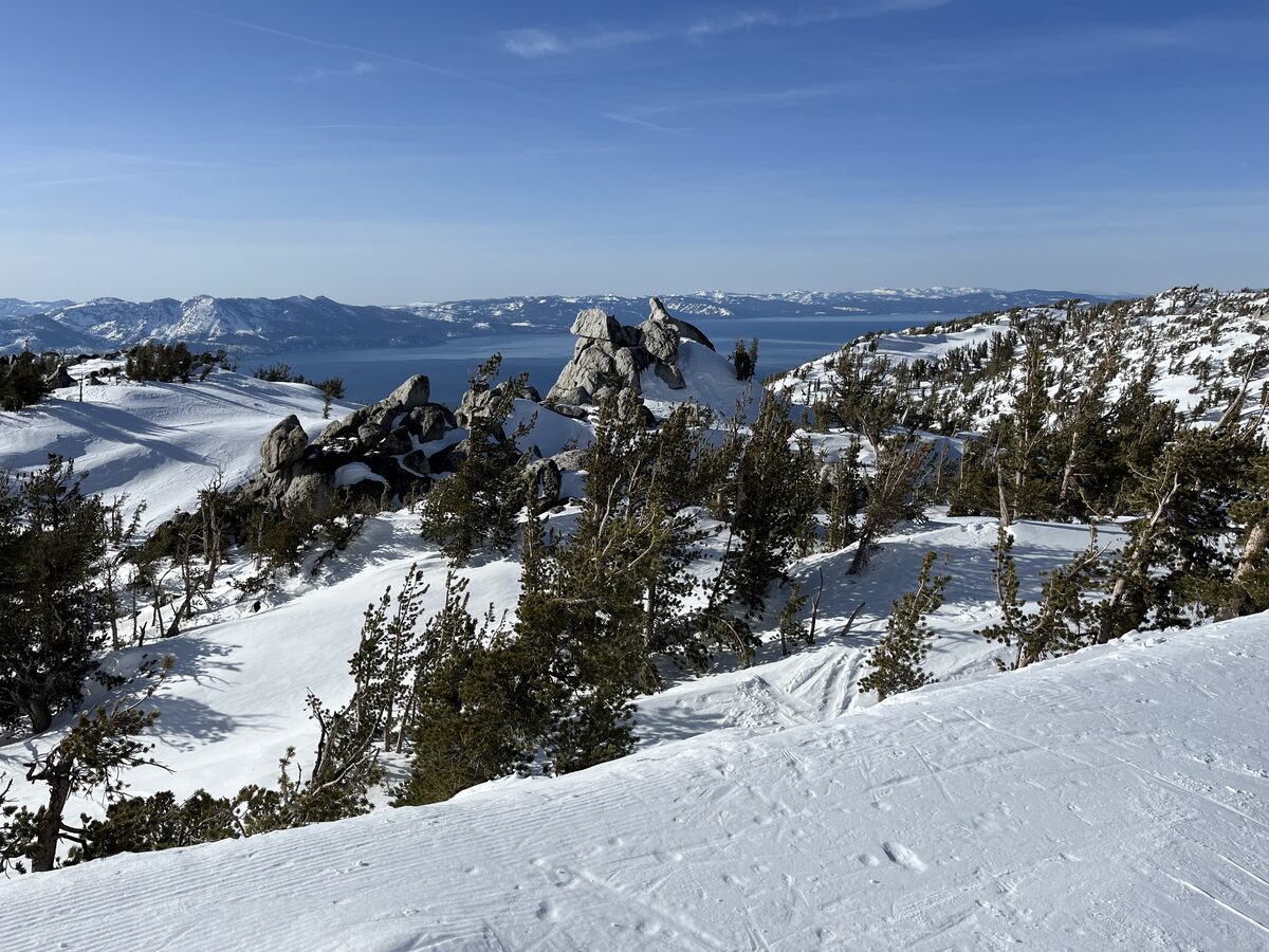 A groomed snowy run above Heavenly, with rock outcrops, pines and Lake Tahoe in the distance