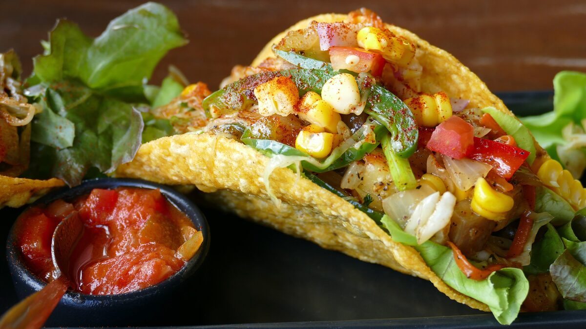 A crisp taco filled with corn, peppers and lettuce, with a small bowl of salsa beside it