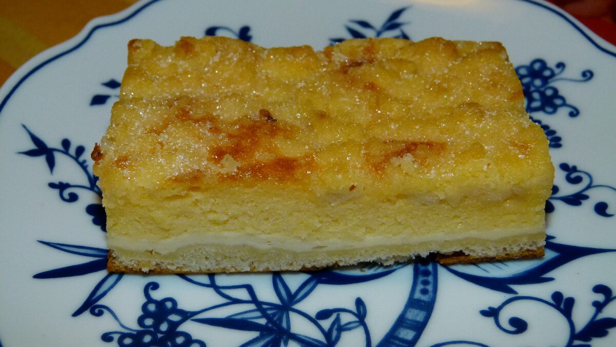 A slice of Eierschecke, with a golden egg custard layer on top of quark filling and a thin cake base, on a blue-and-white plate