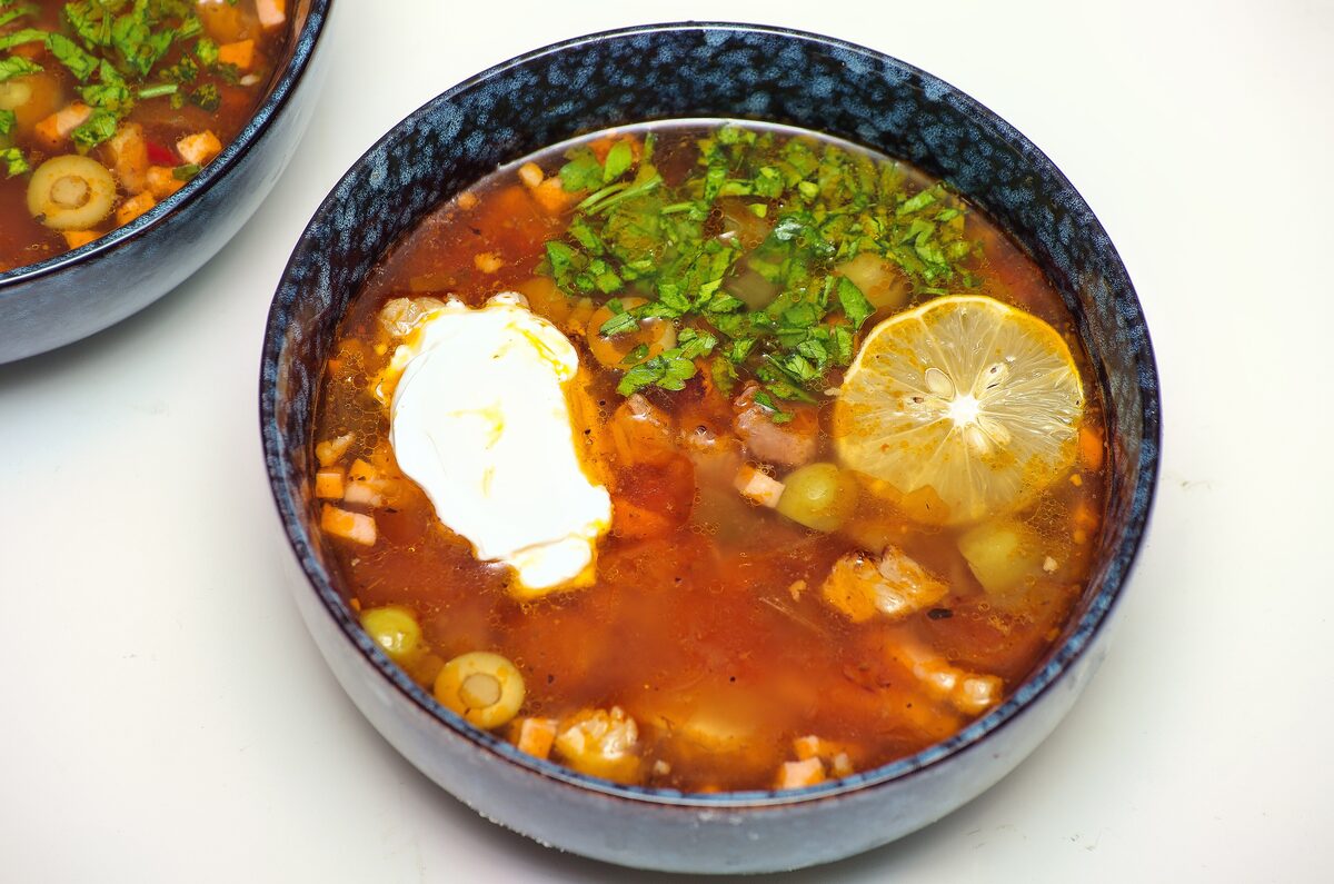 A bowl of soljanka with sour cream, a lemon slice, olives and sausage pieces