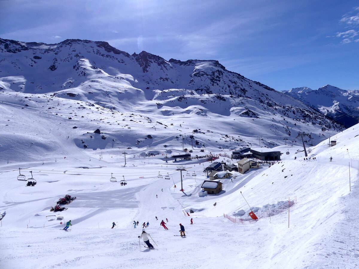 Skiers on the open slopes at Plan Bouchet on the Orelle side, with lifts and snowy peaks behind