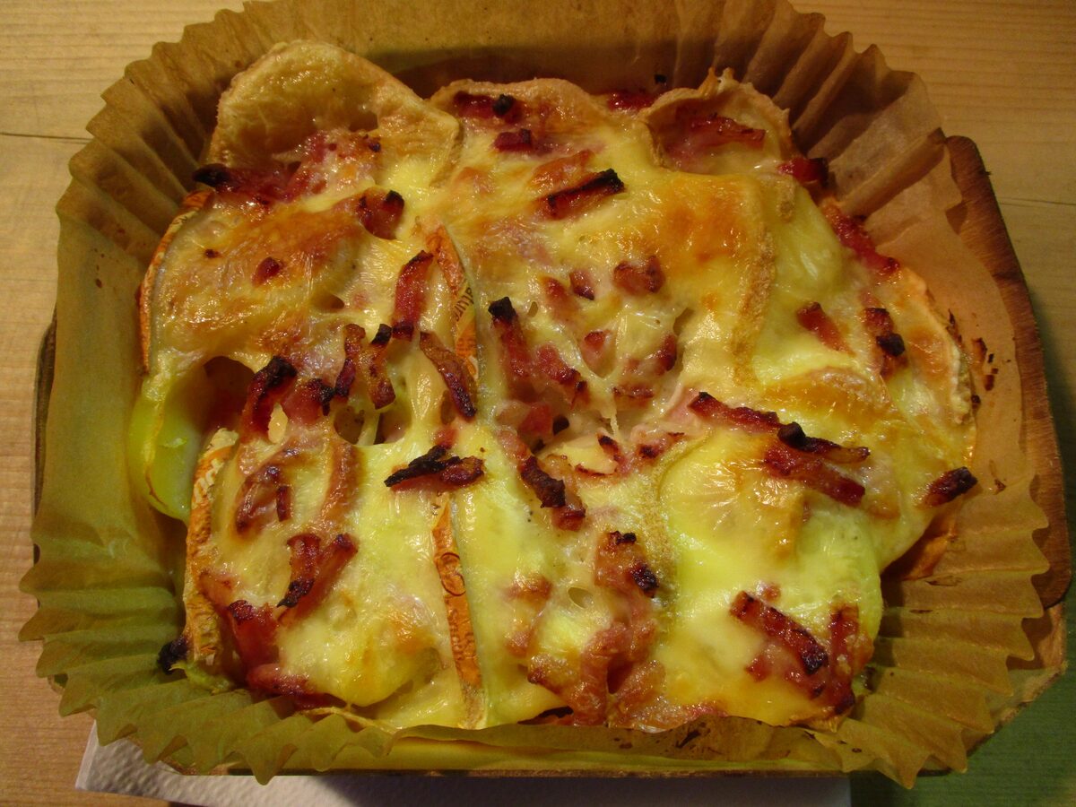 A takeaway portion of tartiflette in a paper tray, potato slices and bacon under browned melted cheese