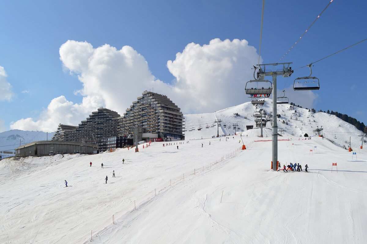 Skiers on the piste below the Paquebot des neiges of Plagne Aime 2000, with a chairlift passing over the slope