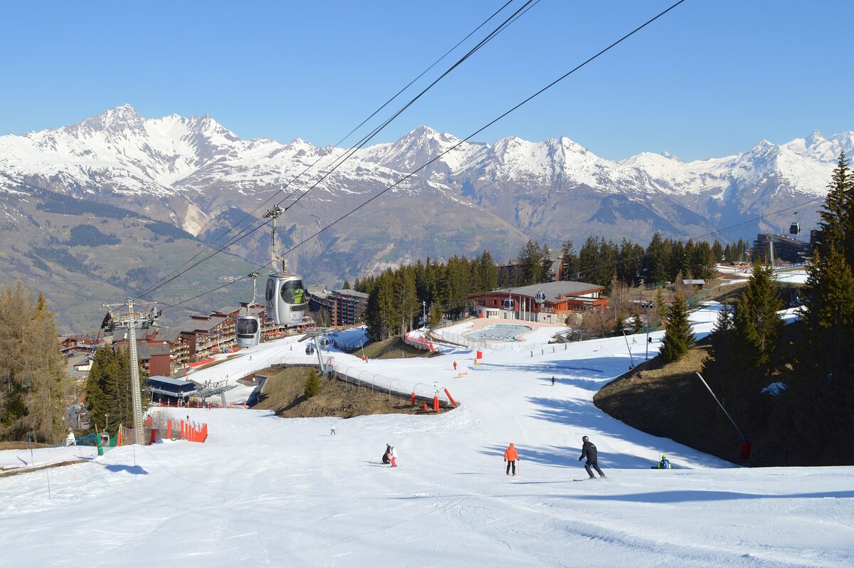 Skiers on the piste into Arc 1800, with a gondola and the snowy Tarentaise peaks behind