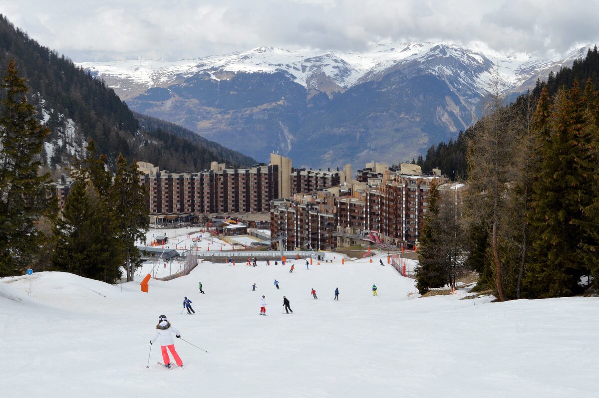 Skiers on the piste in front of the long blocks of Plagne Bellecôte, with snowy mountains beyond the valley