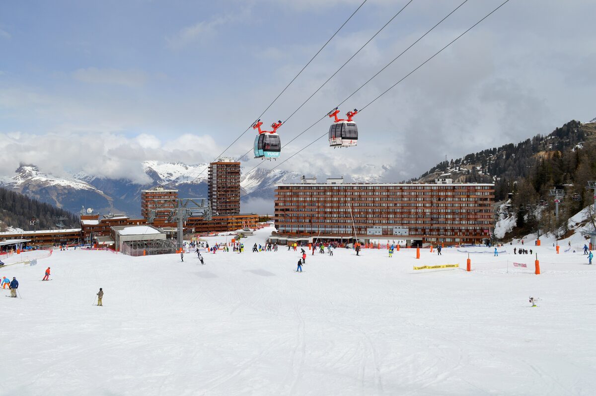 Skiers on the wide snow front of Plagne Centre, with Télémétro cabins running over the slope toward the apartment blocks