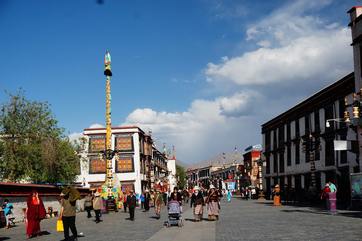 Pedestrians on the paved Barkhor circuit in the old town, with a tall colorful pole and white and black buildings under blue sky