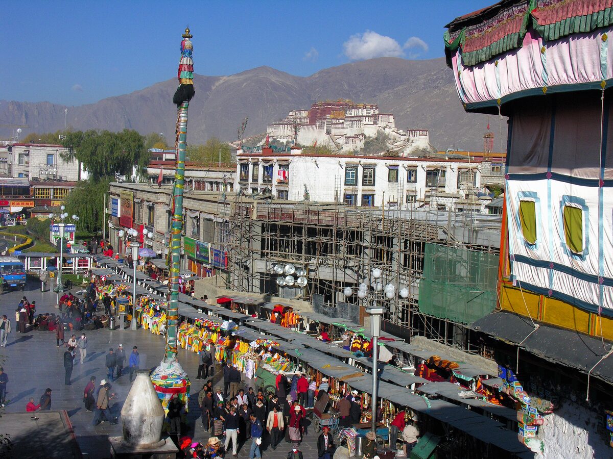 Barkhor Street seen from above, lined with stalls and shoppers, with the Potala Palace on its hill in the distance
